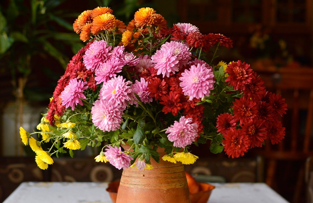 A clay vase holds a colorful bouquet with pink, red, yellow, and orange flowers on a white table