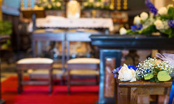 A small bouquet of white and blue flowers rests on a wooden pew, within a decorated church interior featuring red carpet and floral arrangements on the altar.