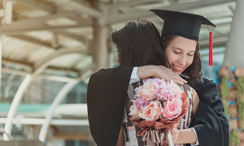 A graduate in cap and gown hugs another person outdoors, holding a bouquet of flowers, under a modern glass and metal structure.