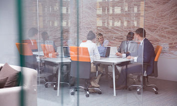 People sitting around a table engaged in discussion in a glass-walled office with modern décor and orange chairs, surrounding a few laptops. An abstract mural covers one wall.