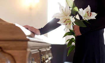 A person in a dark outfit holding white lilies places a hand on a wooden casket in a softly lit room, suggesting a funeral or memorial service.