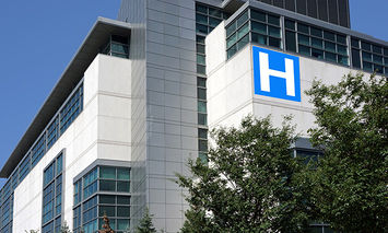 A large, modern hospital building displays a blue sign with a white 