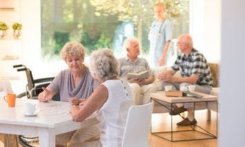 Elderly individualsare conversing at a table inside a well-lit room with large windows, while a nurse stands near another seated group