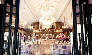 A grand ballroom, adorned with chandeliers and elegant floral arrangements, is set for a formal event with round tables, white chairs, and ornate decorations.